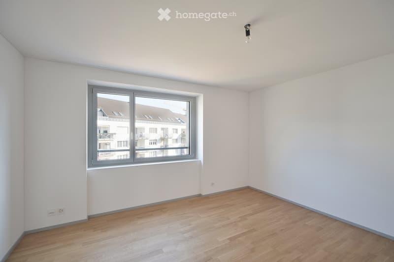Empty room with light wooden flooring, white walls, and a large window showing a view of neighboring buildings.