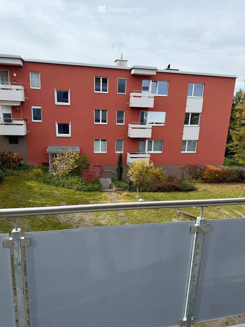 Red apartment building with multiple balconies and windows, surrounded by a small garden area with grass and shrubs.