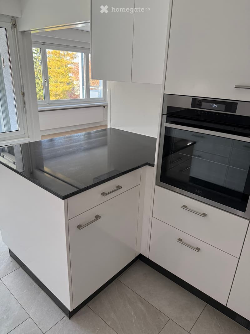 Modern kitchen with white cabinets, black countertop, and built-in oven. Large window provides natural light.