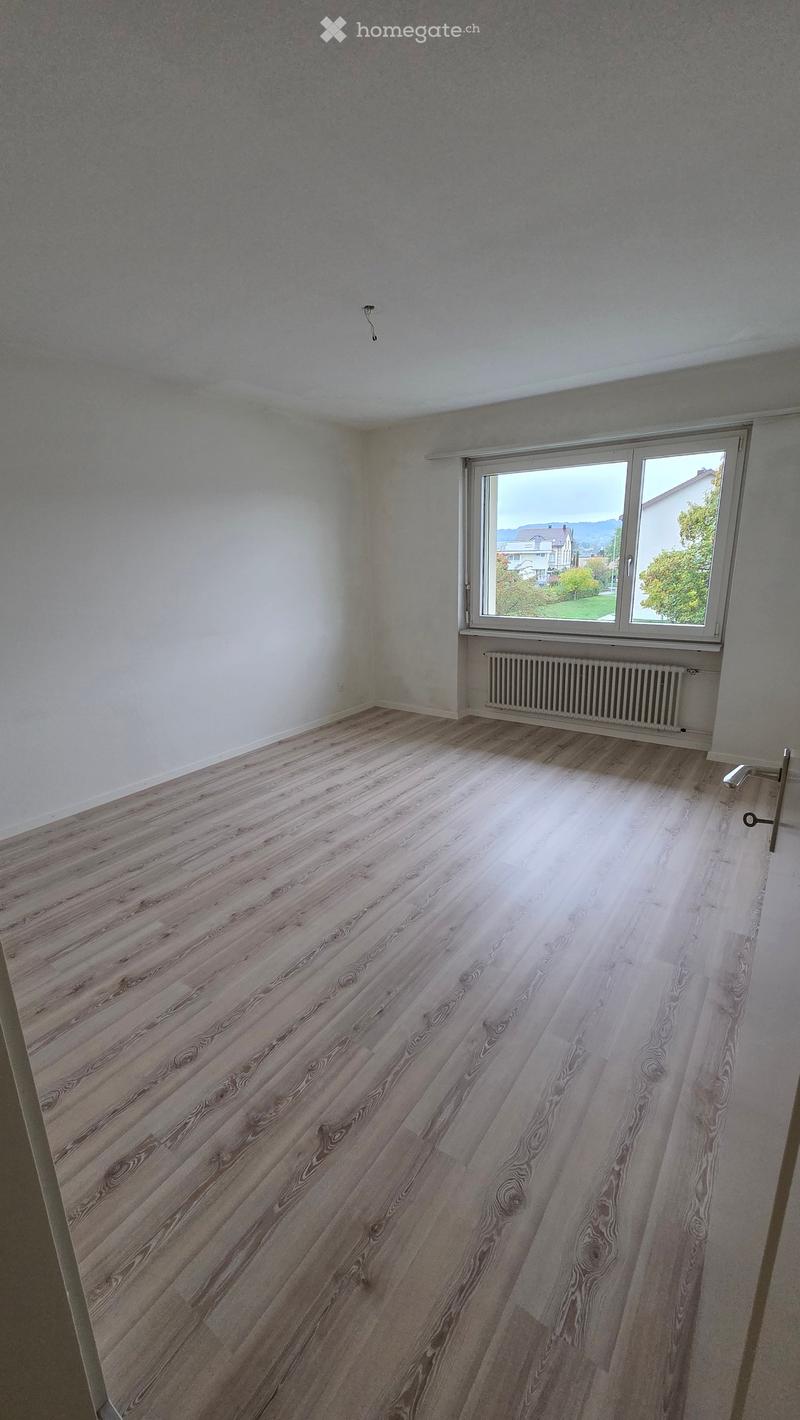 Empty room with light wooden flooring, white walls, and a large window with a view of the outside. A radiator is visible beneath the window.