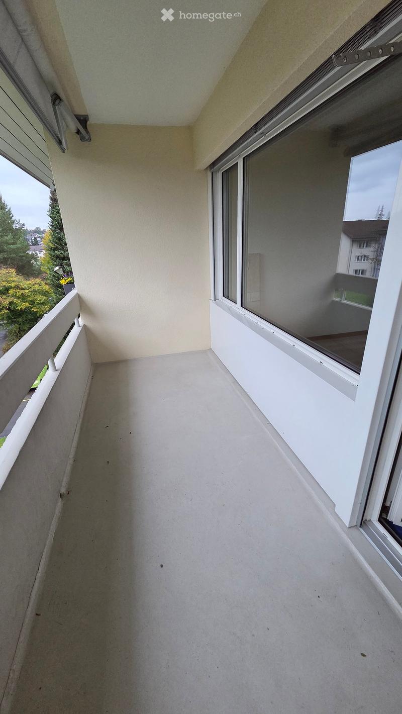 Empty balcony with white railing, large window, and view of greenery and neighboring building.