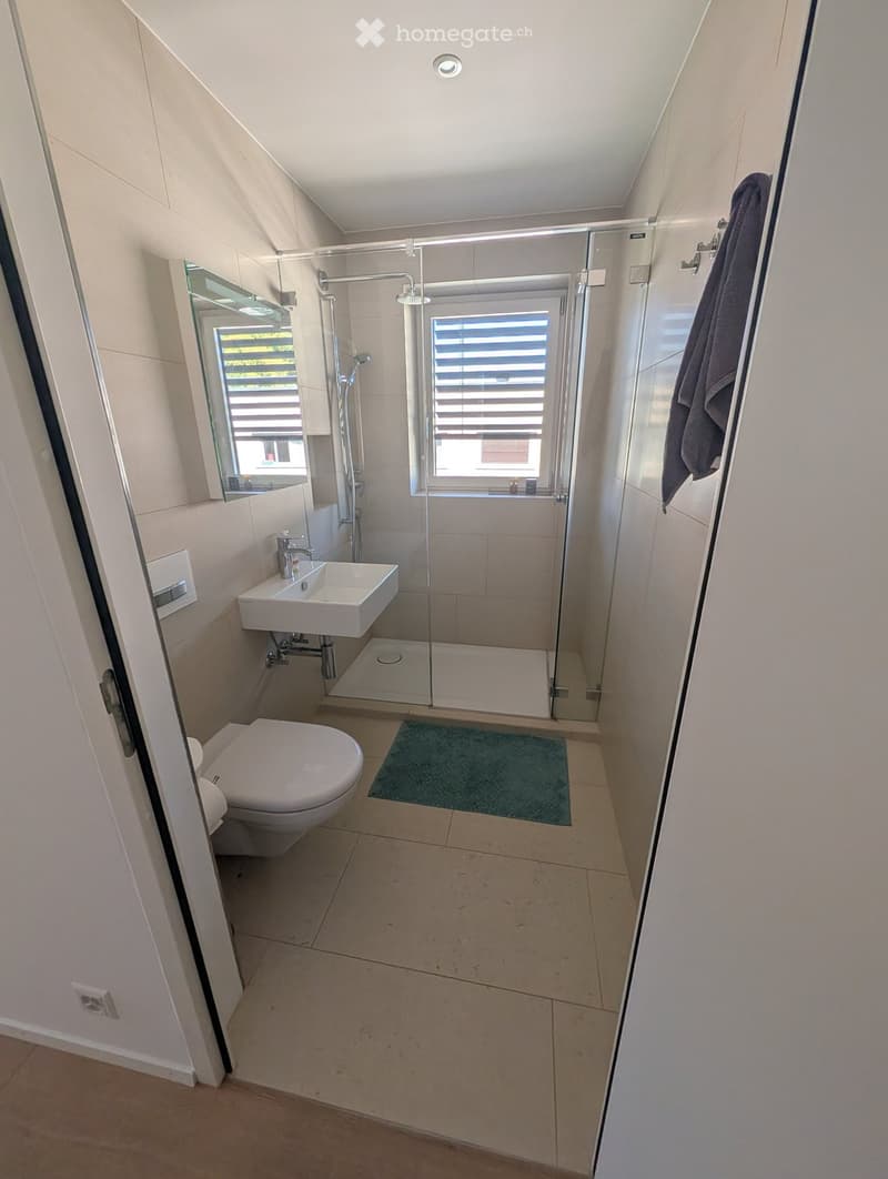 Modern bathroom with a toilet, sink, shower, and a towel hanging on the wall. The space features light-colored tiles and a small window with blinds.
