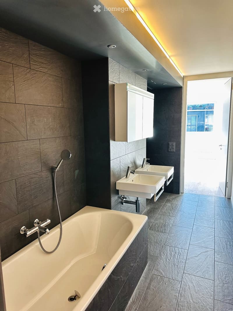 Modern bathroom with a bathtub, two sinks, a mirror, and a sleek cabinet. The space features dark grey tiles and a warm, recessed lighting system.