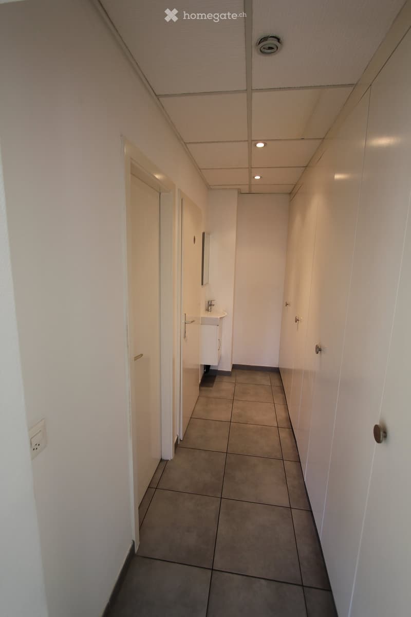 A narrow hallway with white walls, tiled floor, and several doors leading to rooms. A small bathroom is visible at the end of the hallway, featuring a sink, mirror, and cabinet.