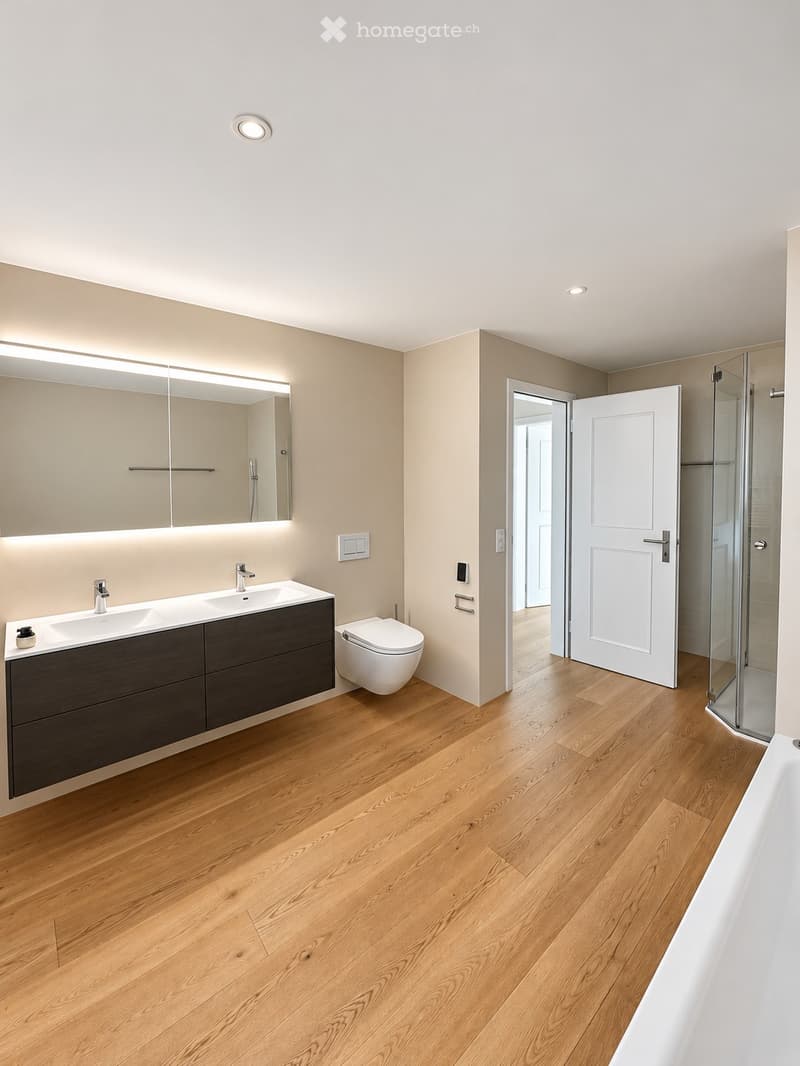 Modern bathroom with light wood flooring, double sink vanity, toilet, large mirror, and glass shower enclosure.