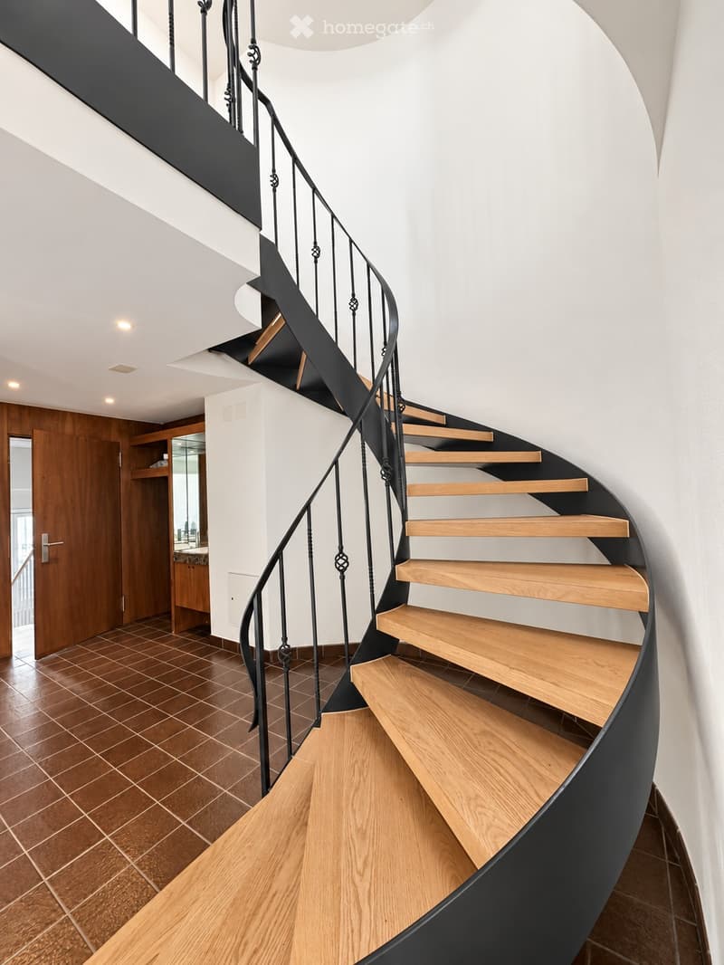 Spiral staircase with wooden steps and black wrought iron railing, leading to an upper level. The hallway features tiled flooring and white walls.
