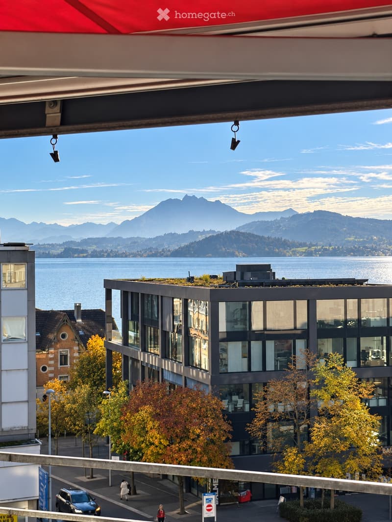 Modern multi-story building with large glass windows, surrounded by autumn trees and overlooking a lake and mountain range.