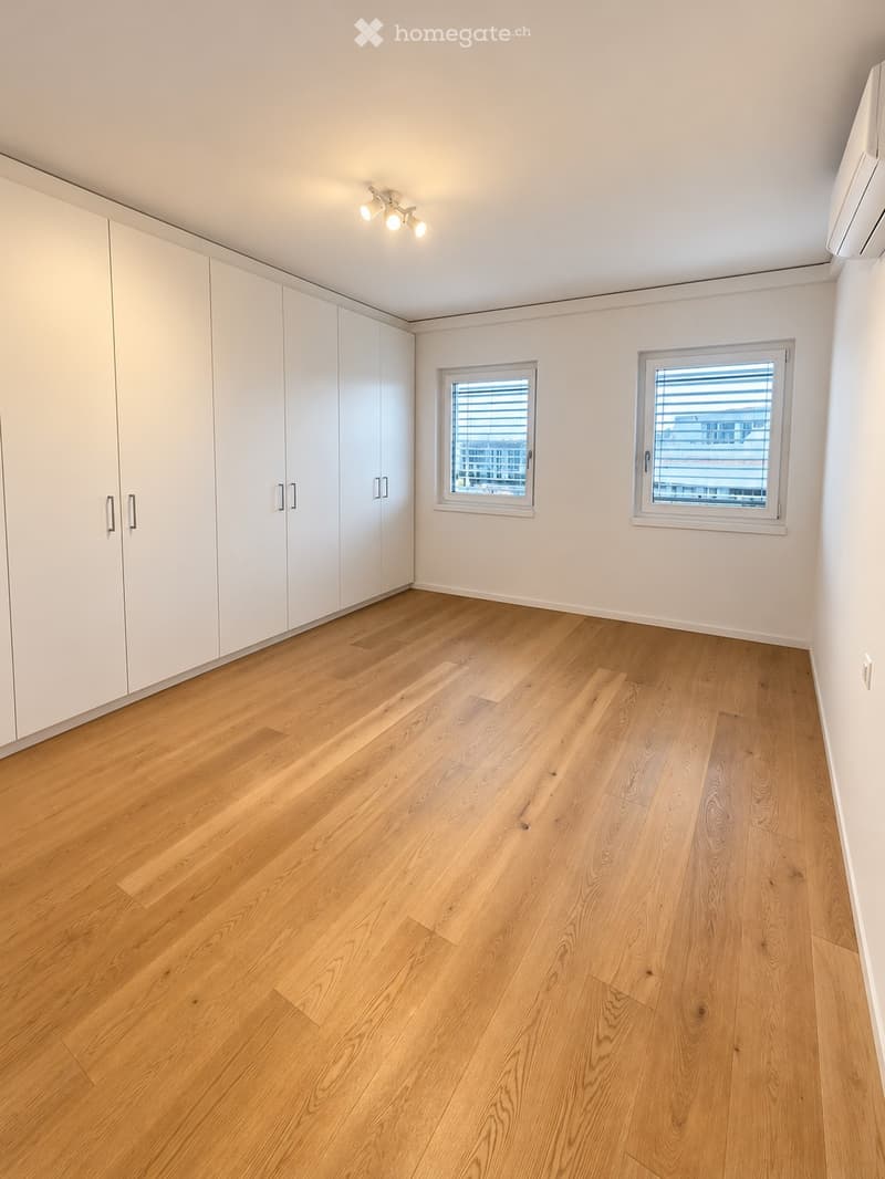 Empty room with light wooden flooring, white walls, built-in wardrobes, and two windows with blinds.
