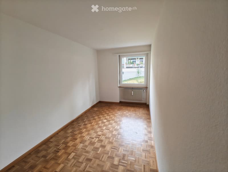 Empty room with wooden herringbone flooring, white walls, and a window with a radiator below it.