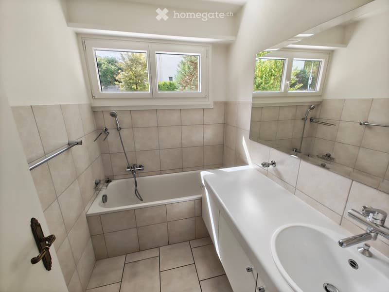 Modern bathroom with a bathtub, sink, mirror, and showerhead. Features tiled walls and a large window letting in natural light.