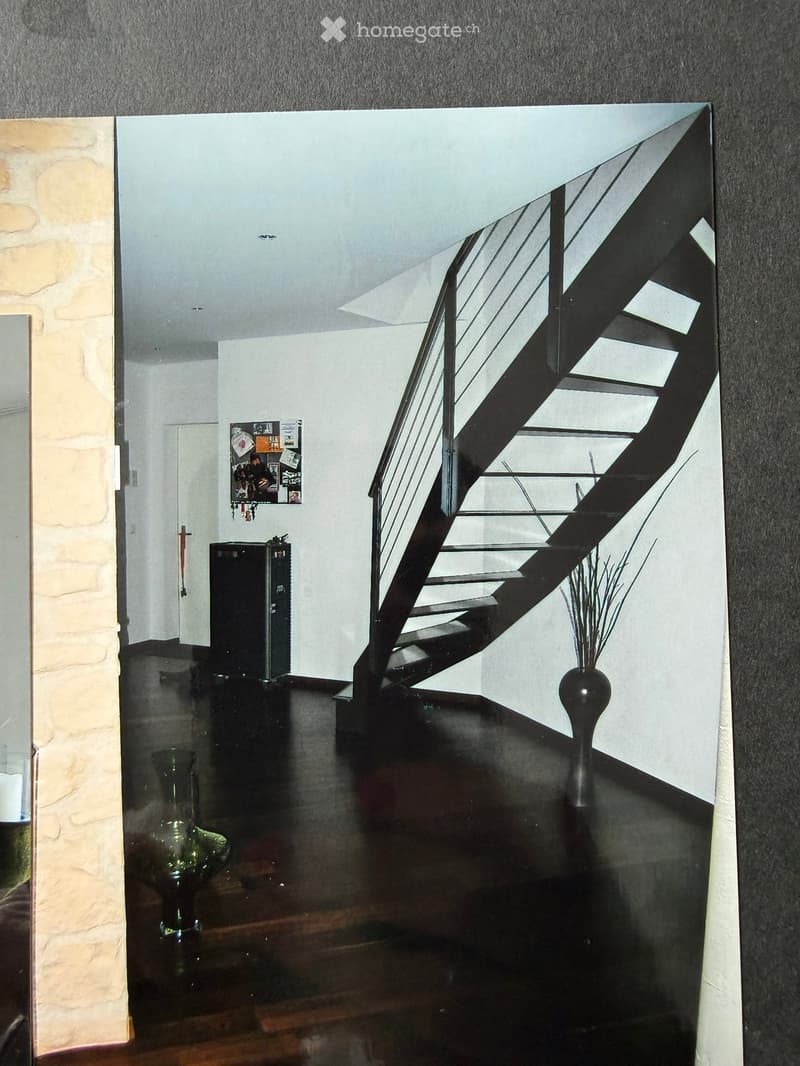 Modern hallway with a staircase featuring a glass railing, wooden steps, and a black metal balustrade. The walls are painted in neutral tones, and there are two vases placed on either side of the staircase.
