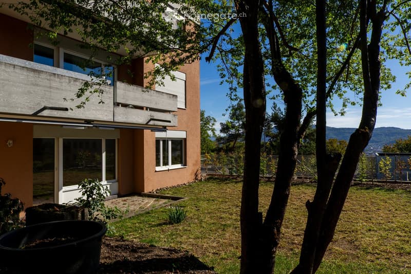 Garden area with a modern two-story house in the background, featuring a balcony, large windows, and a scenic mountain view.
