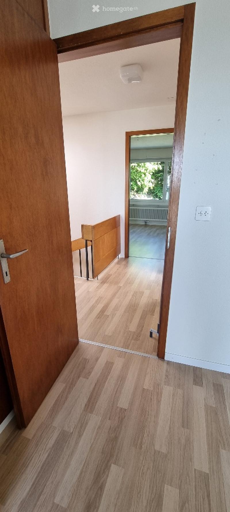 Open wooden hallway with light-colored wooden flooring, white walls, and a visible adjacent room with a radiator and large window.