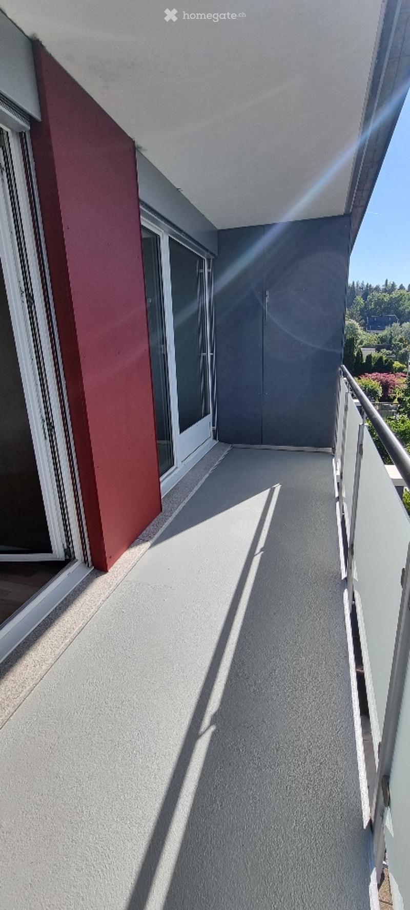 Balcony with grey flooring, white railing, large windows, and a view of greenery.