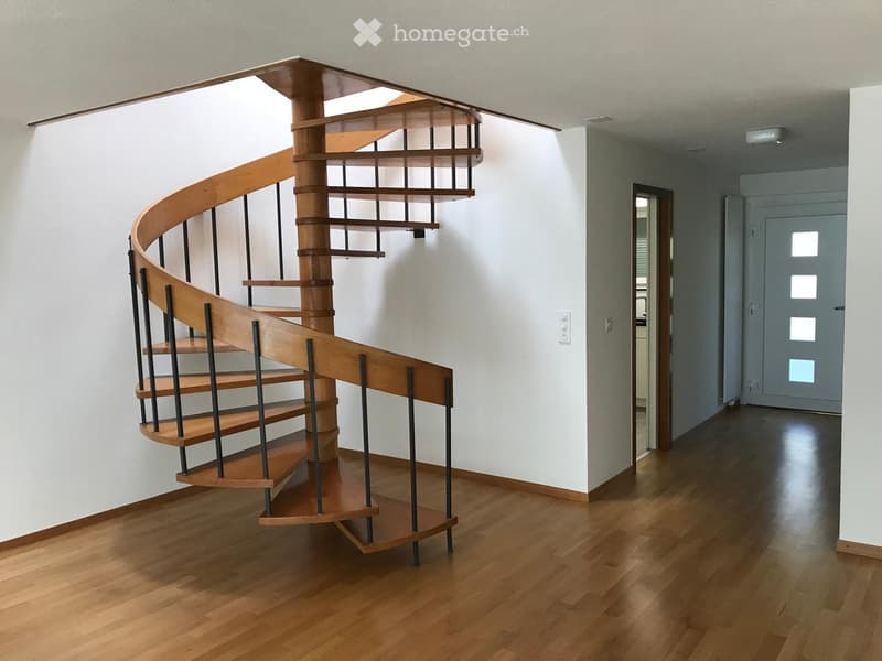 Spiral staircase with wooden steps and metal railings, light-colored walls, polished wooden floor, and a closed door at the end of the hallway.