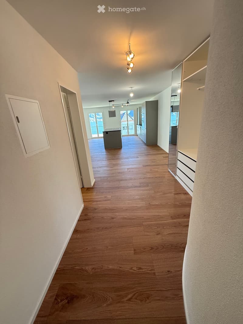 Empty hallway with wooden flooring, white walls, and built-in storage on one side. The hallway leads to a room with a kitchen area visible in the background.