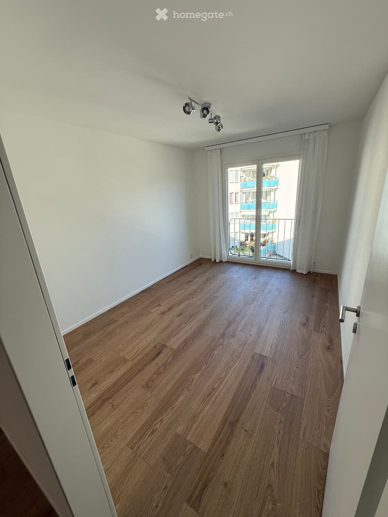 Empty room with wooden flooring, white walls, large sliding glass door with white curtains, and track lighting fixture on the ceiling.
