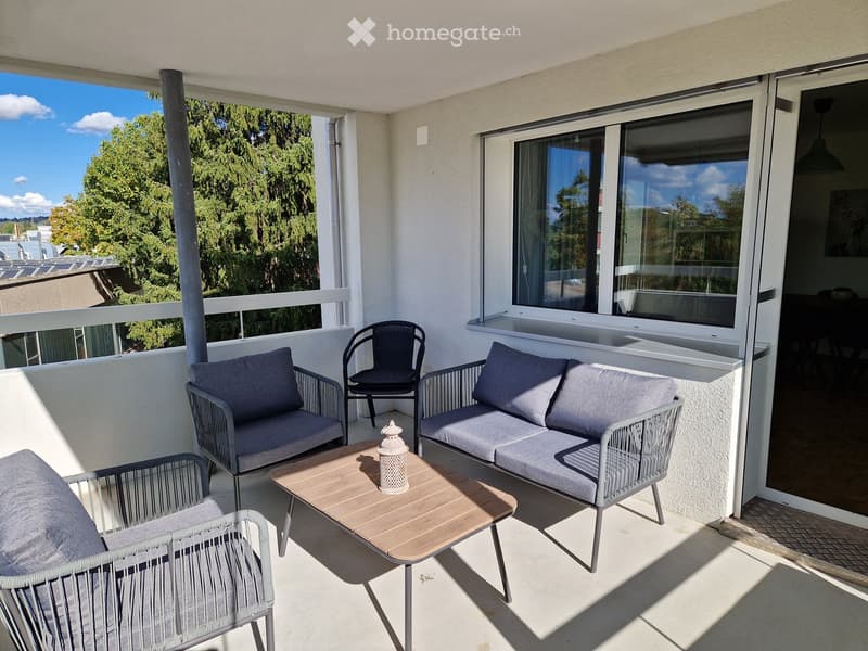 Balcony with a sofa, chairs, a wooden coffee table, and cushions, overlooking a tree-lined area.