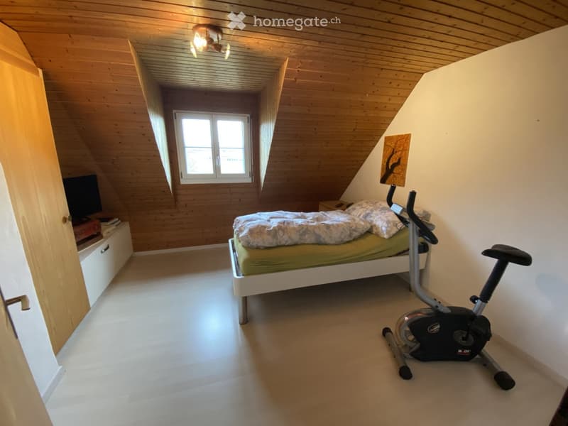 Cozy attic bedroom with wooden slanted ceilings, a single bed, exercise bike, nightstand, and a TV screen on a stand.