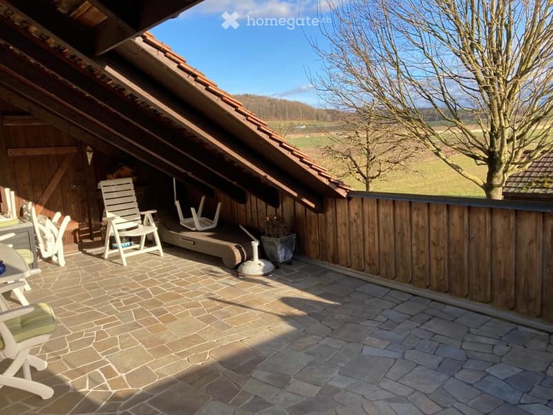Wooden balcony with stone tiles, white chairs, small table, and potted plant, overlooking a rural landscape.