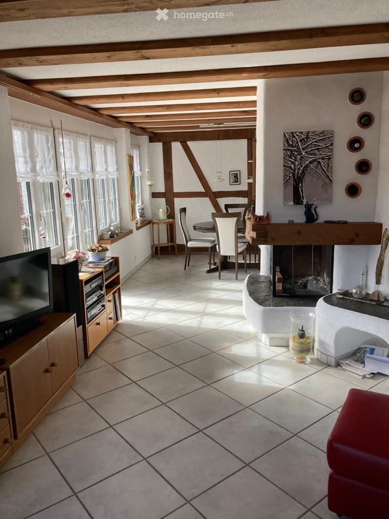 A cozy living room with exposed wooden beams, a fireplace, a television, a dining area with chairs and a table, and a small seating area with a red chair and a vase on a table.