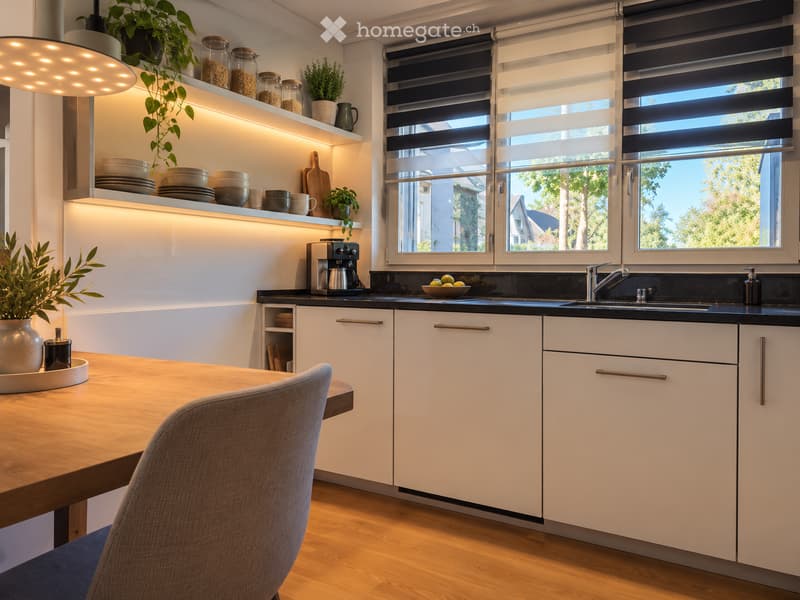 Modern kitchen with white cabinets, wooden table, grey chair, sink, coffee maker, and decorative jars on shelves.