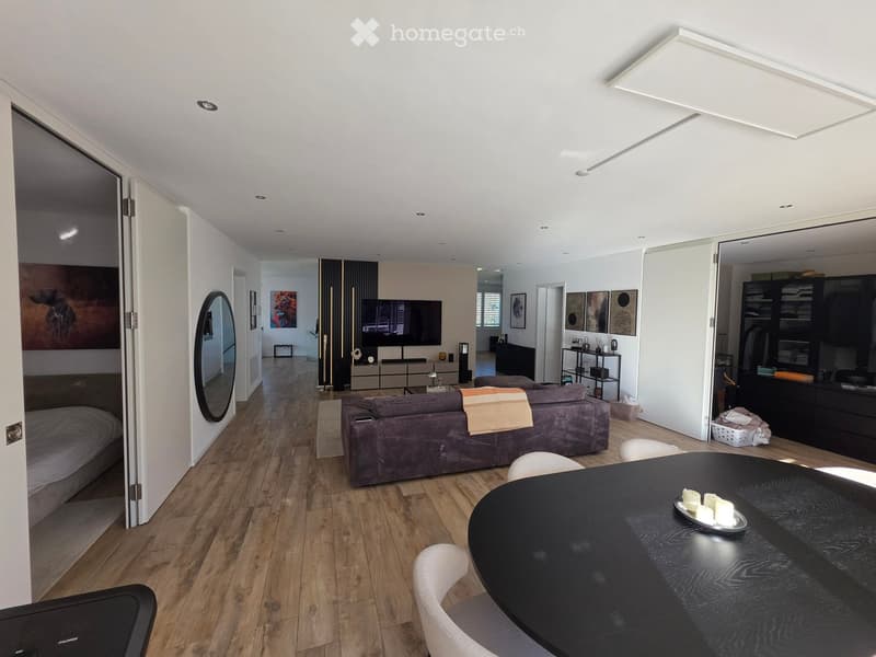Modern, open-plan living and dining area with a grey sofa, black dining table, white chairs, a round mirror, and a TV in the background. The space features wooden flooring and white walls.