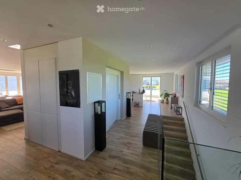Modern hallway with wooden flooring, white walls, large windows, and a staircase leading to another level. Features include a glass railing, a bench, and a large artwork on the wall.