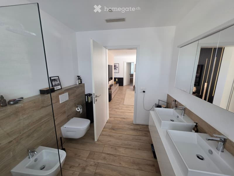 Modern bathroom with wooden flooring, white walls, two sinks, a toilet, a bidet, and a large mirror. A framed picture and a bottle are on the wooden vanity.