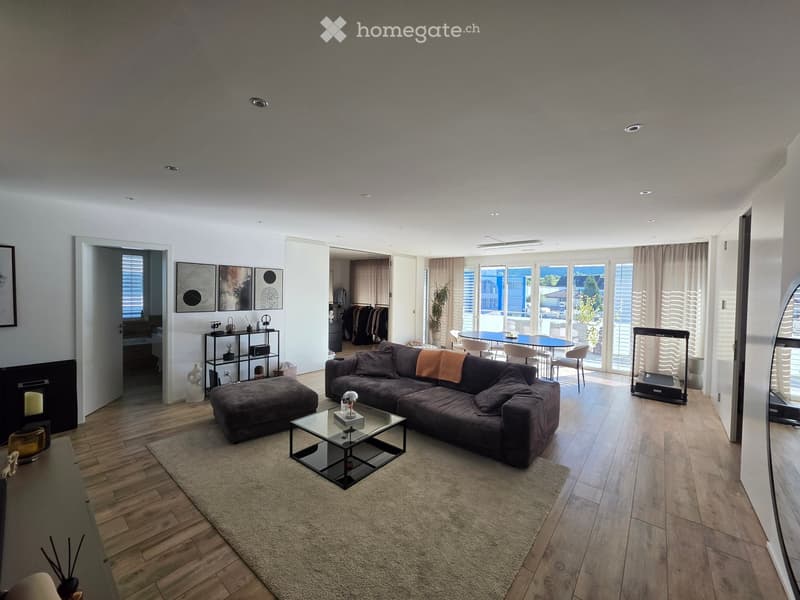 Modern living room with a large sectional sofa, glass coffee table, ottoman, and a dining area with a table and chairs. Features a fireplace, wooden flooring, and large windows with a view.