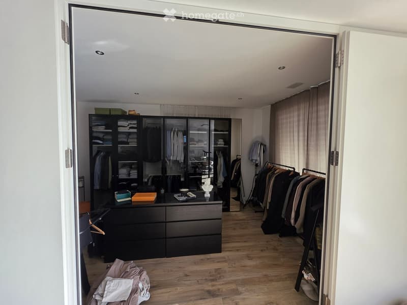 A well-organized walk-in closet with wooden flooring, large wardrobe with mirrored doors, a dresser, and a clothing rack with hanging garments. The space includes a small table with books, a hat, and a decorative statue.