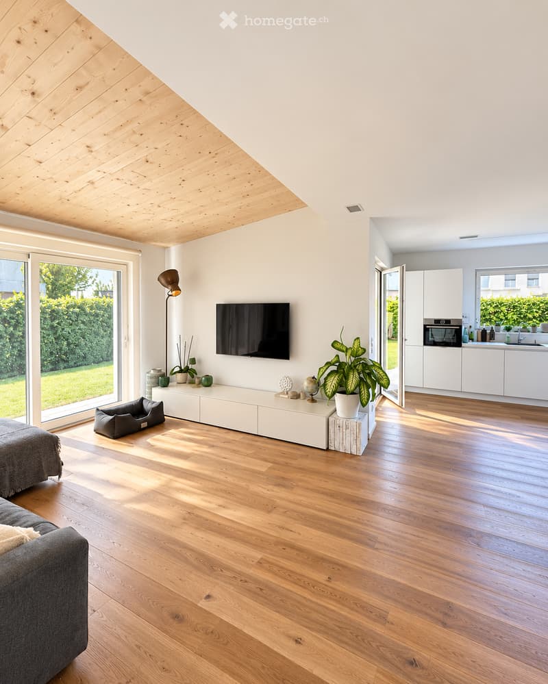 Modern living room with wooden flooring, grey sofa, TV mounted on wall, floor lamp, console table with decorative items, pet bed, and an open kitchen area with white cabinets and appliances.