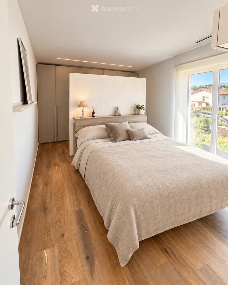 Modern bedroom with wooden flooring, large bed with beige bedding, nightstand with lamp, potted plant, and built-in wardrobe.
