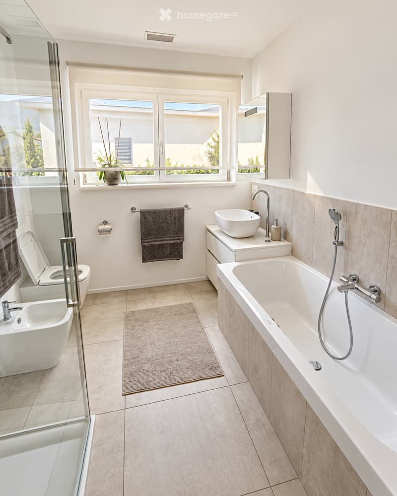 Modern bathroom with a bathtub, toilet, sink, and shower area. Features include a large window with a view, a towel rack, a mirror, and a small plant on the windowsill.