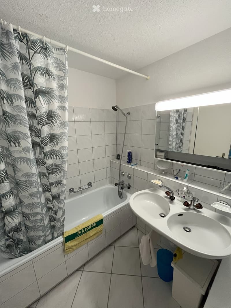 Bathroom with a bathtub, double sink, mirror, and shower area. Features a palm leaf patterned shower curtain and a yellow towel on the edge of the tub.