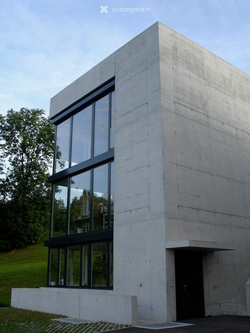 Modern, minimalist concrete building with large glass windows and a raised ground floor entrance.