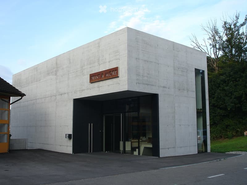 Modern concrete building with a sign reading "PRINT & MORE" above the entrance, featuring large glass windows and doors.