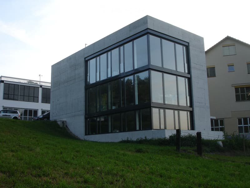 Modern concrete building with large glass windows, situated on a raised ground floor with a grassy slope leading up to it. Adjacent buildings and parked cars are visible in the background.