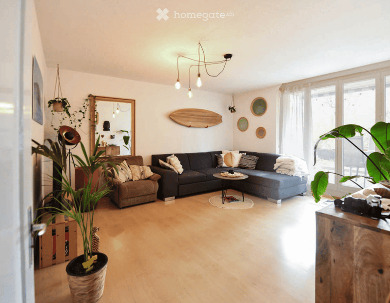 Modern living room with light wooden flooring, large windows, a dark blue sectional sofa, a small round coffee table, and several potted plants. The room features a hanging chandelier, a large mirror, and decorative wall art.