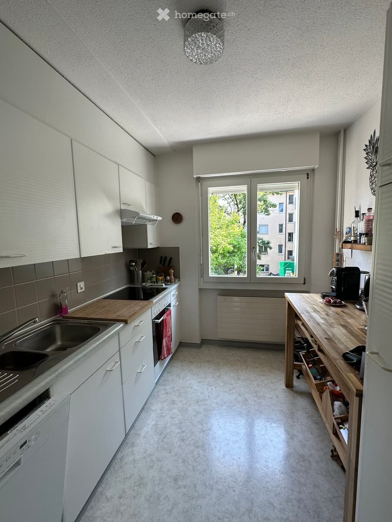 Modern kitchen with white cabinets, wooden countertop, sink, oven, dishwasher, and various appliances. Features a window with a view of greenery and a city building.