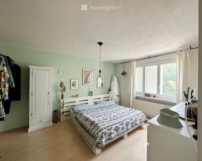 A modern bedroom with a white wooden bed, geometric patterned bedspread, white wardrobe, nightstand, surfboard, and various decorative items including picture frames and candles.