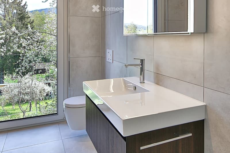 Modern bathroom with a white sink, sleek faucet, toilet, and wooden cabinet, large window offering a scenic view of blooming trees and greenery.