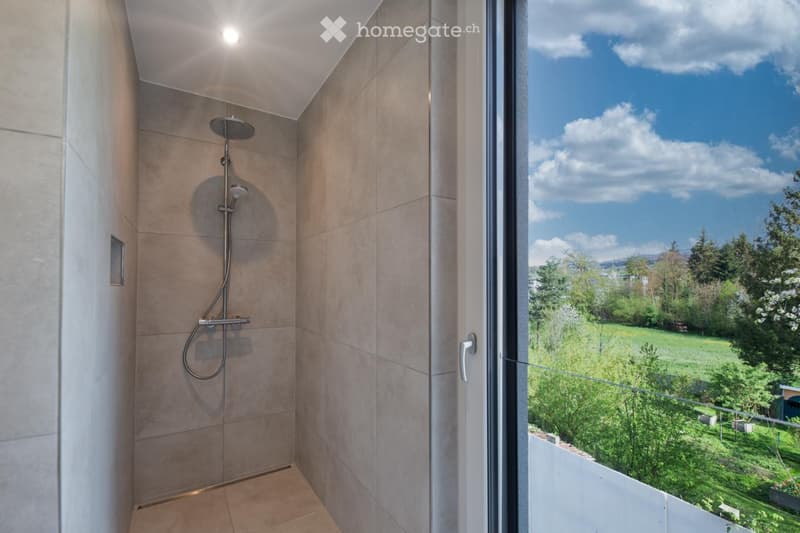 Modern bathroom with a large window offering a scenic view of a green landscape and mountains. Features a sleek shower area with a modern showerhead and handle.