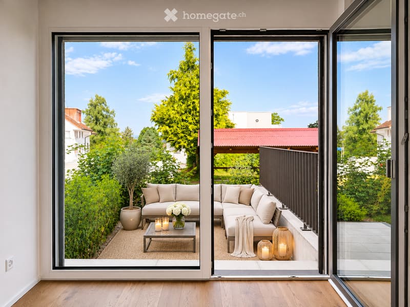 Modern balcony with large glass doors, beige sofa, coffee table, candles, and potted plant, overlooking a green garden.