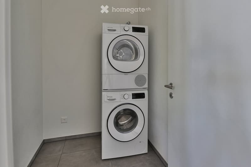 A compact room with a stacked washing machine and dryer unit, white walls, tiled floor, and a door.