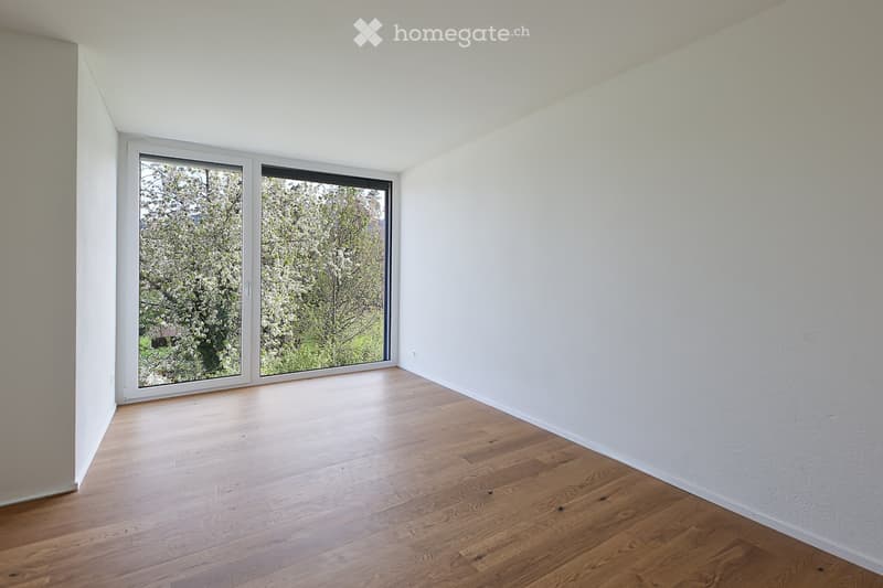 Empty room with light wooden flooring and white walls, featuring large windows with a view of trees and greenery outside.