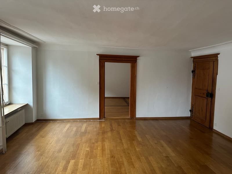 Empty room with wooden flooring, light-colored walls, and a large window on one side. There is a radiator below the window and a wooden door leading to another room.