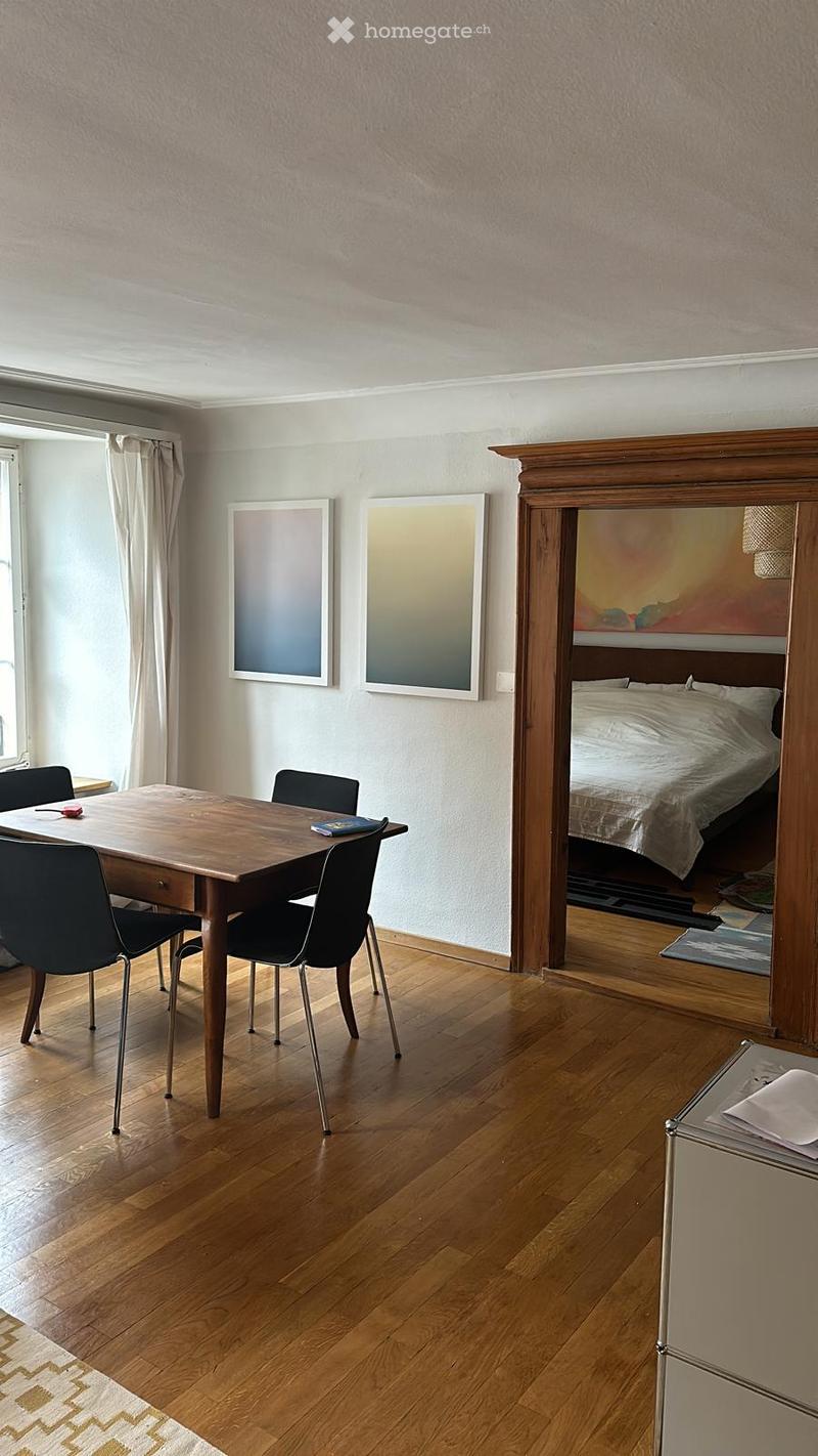 A modern dining area with a wooden dining table and chairs, adjacent to a bedroom with a bed and artwork on the walls.