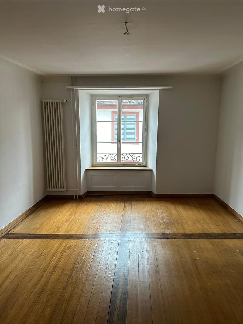 Empty room with wooden flooring, white walls, a window with shutters, and a radiator on the left side.