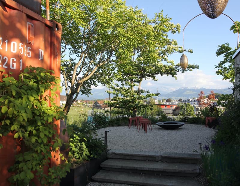 Garden area with gravel pathway, steps, red stools, fire bowl, and hanging decorative light, surrounded by greenery and a view of mountains.